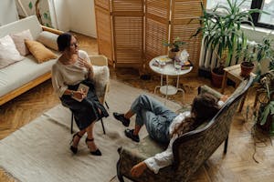 Two women engaged in a psychotherapy session in a warm, inviting interior with plants and natural lighting.
