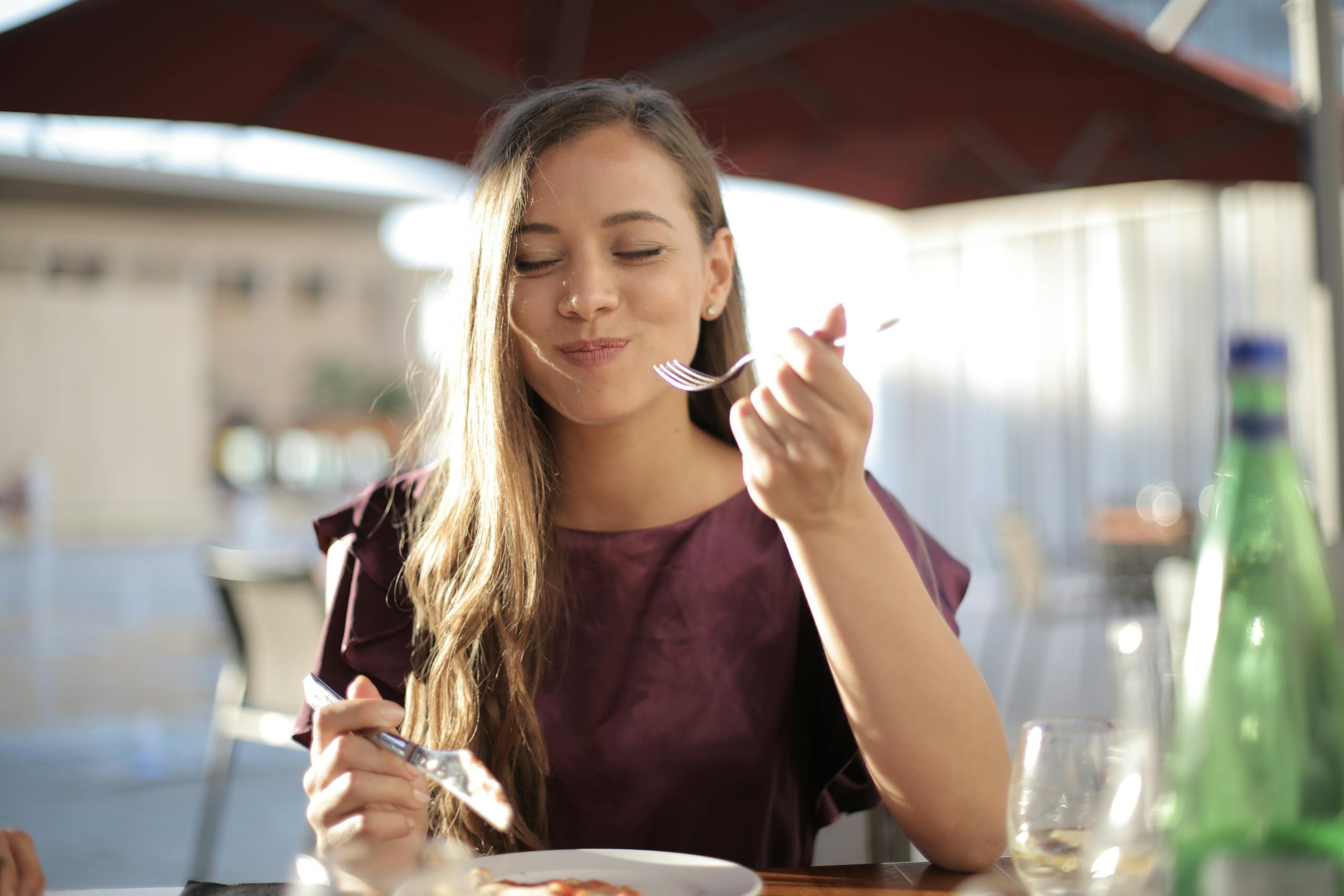 A woman savoring her meal, eating, with a smile, dining outdoors in a pleasant setting.