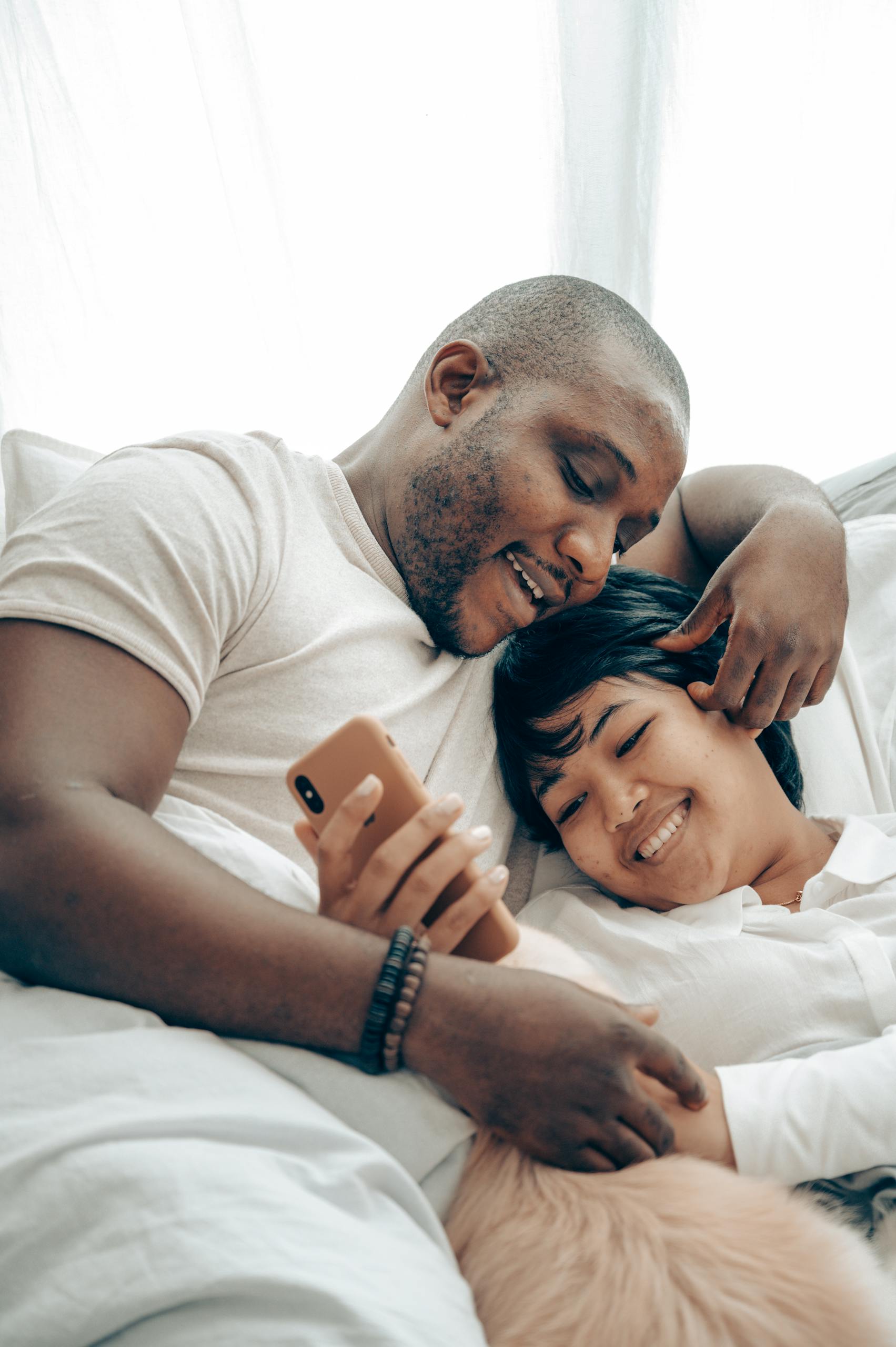 Cheerful young diverse couple lying together in bed and smiling while watching video on mobile phone in morning