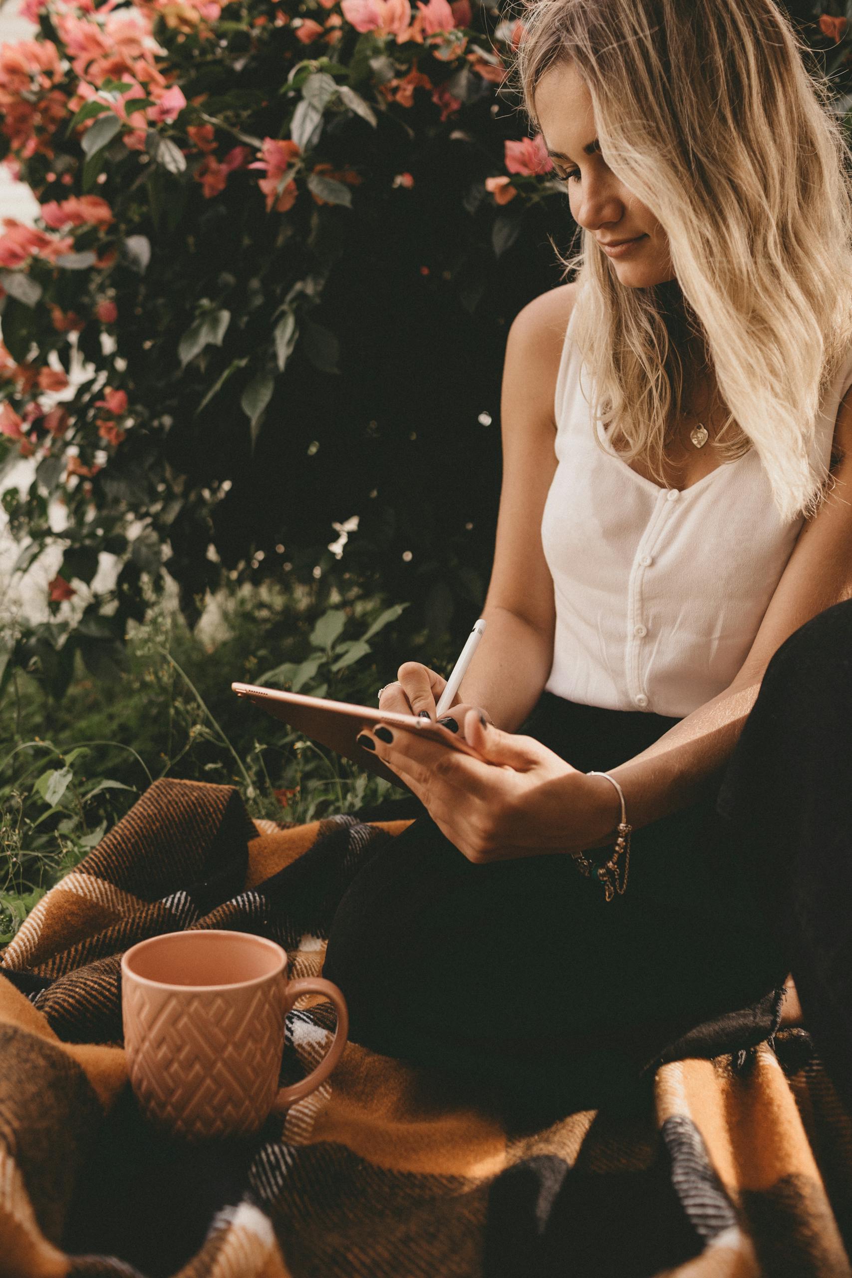 Young woman writing on tablet while relaxing outdoors with a coffee, surrounded by flowers.